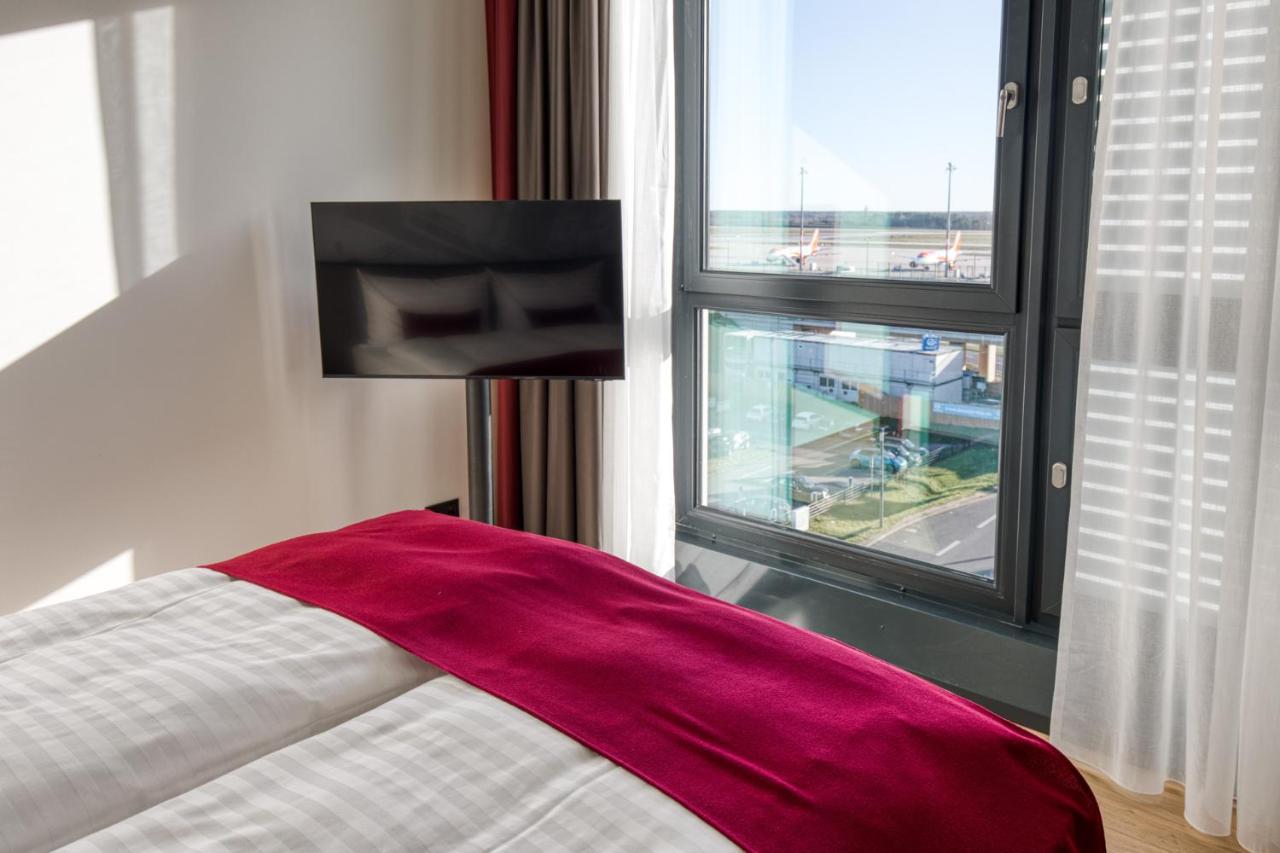 Intercityhotel Berlin Airport Ber Terminal 1 & 2 Шёнефельд Экстерьер фото The photo shows a modern hotel room with a large bed covered in white linens and a burgundy throw blanket. In the background, there is a large window that lets in natural light, offering a view of the outside. A flat-screen TV is mounted on the wall 