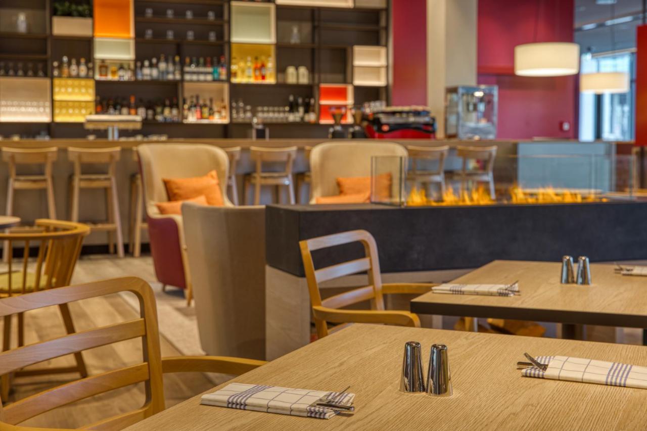 Intercityhotel Berlin Airport Ber Terminal 1 & 2 Шёнефельд Экстерьер фото The photo shows a modern restaurant or bar interior. In the foreground, there are wooden tables set with napkins and condiments. The seating includes wooden chairs and plush seating in various colors in the background. A stylish bar area is visible w